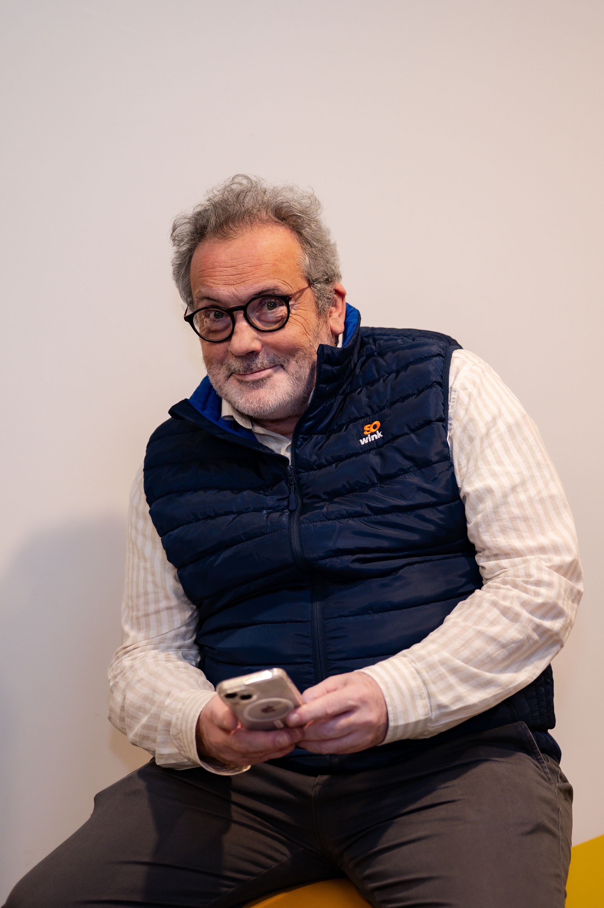 Un homme portant des lunettes, une chemise blanche et un gilet bleu marine, est assis et regarde un smartphone sur un fond blanc uni.