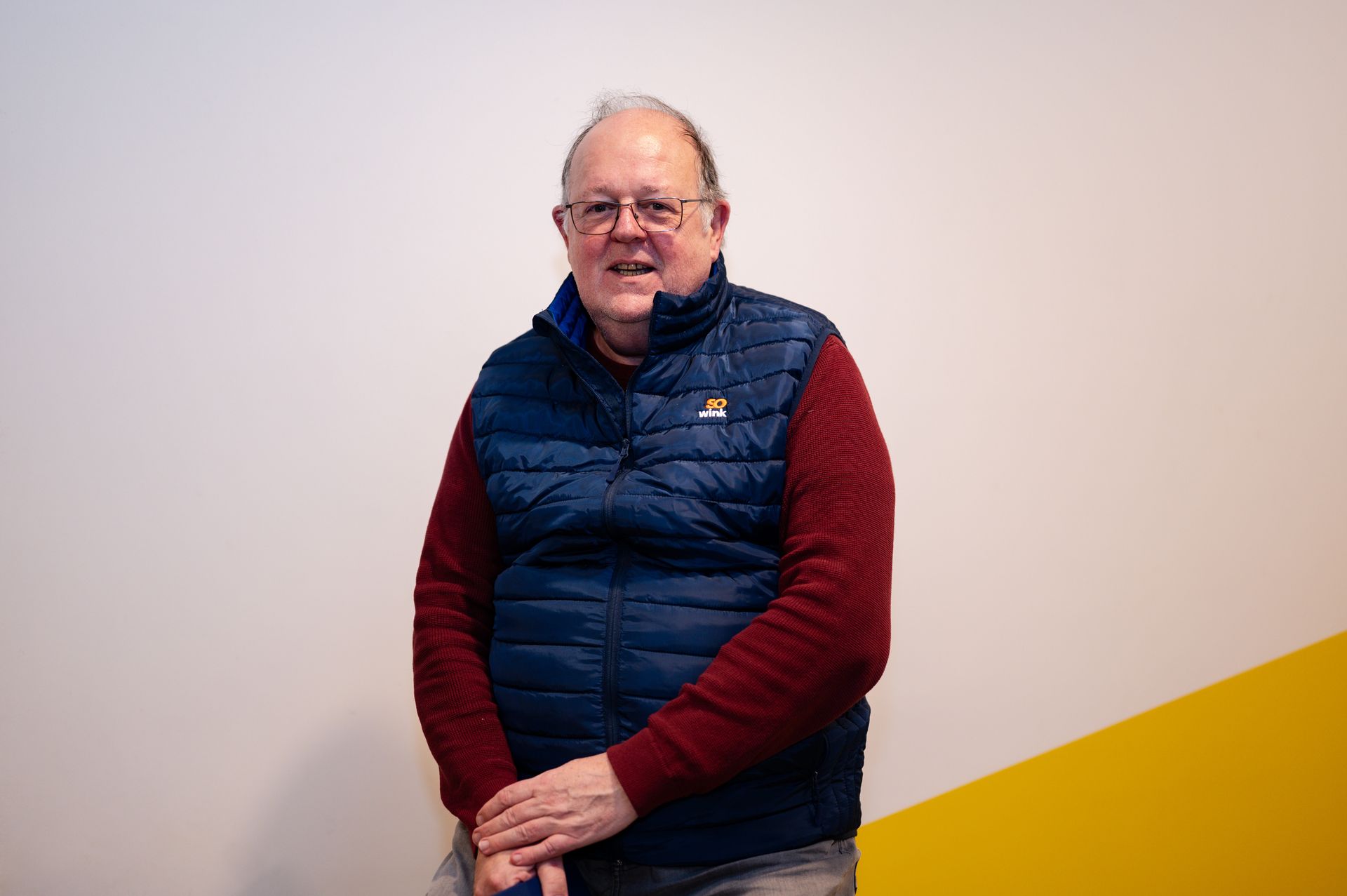 Une personne vêtue d'un pull bordeaux et d'une doudoune sans manches bleue se tient devant un mur blanc et jaune, souriant à l'objectif.