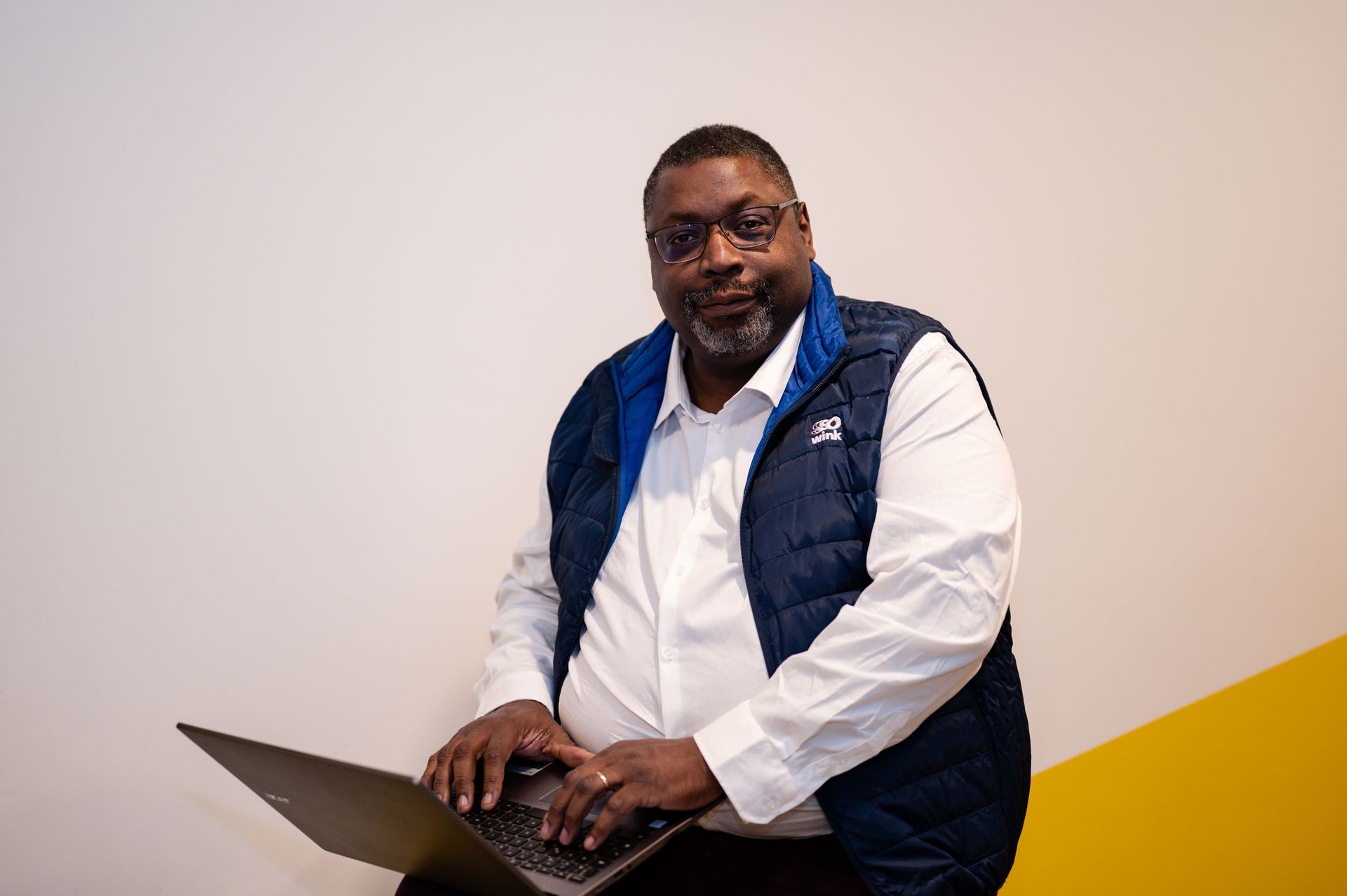 Une personne vêtue d'une chemise blanche et d'un gilet bleu marine est assise, tapant sur un ordinateur portable adossé à un mur blanc et jaune.
