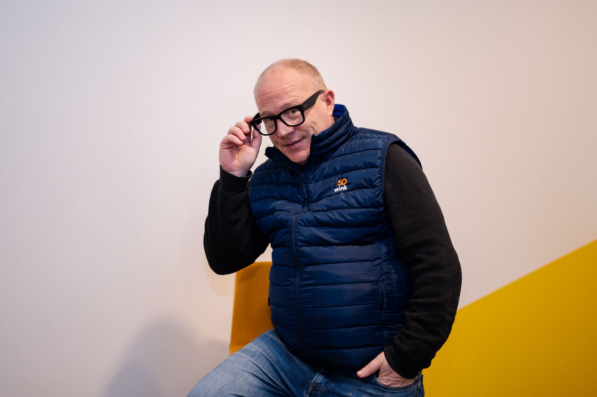 Une personne vêtue d'une doudoune sans manches bleue et d'un pull noir ajuste ses lunettes, assise devant un mur jaune.