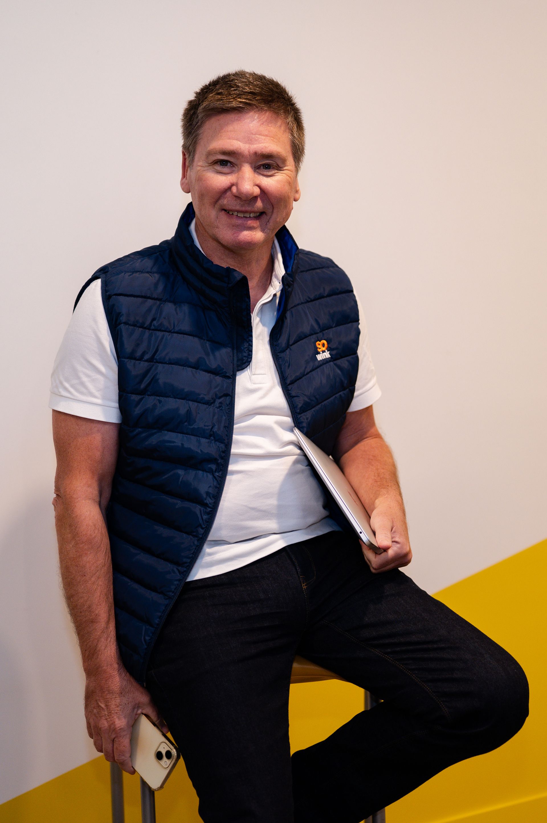 Une personne assise sur un tabouret, vêtue d'une chemise blanche et d'un gilet bleu foncé, tenant un téléphone et un ordinateur portable, adossée à un mur.