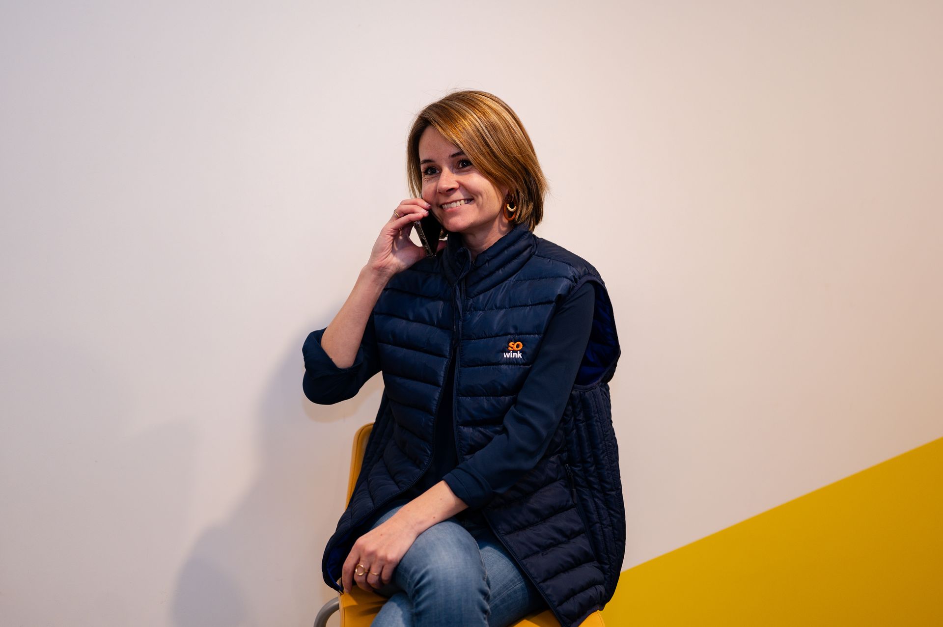 Une personne vêtue d'un gilet bleu est assise sur une chaise jaune, adossée à un mur blanc et jaune, souriante tout en parlant au téléphone avec un smartphone.