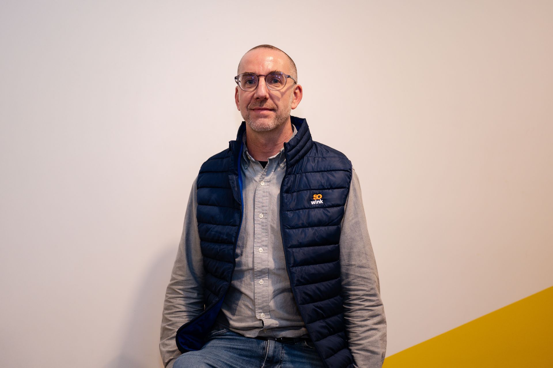 Un homme en chemise boutonnée et gilet matelassé bleu se tient devant un mur blanc avec un coin jaune.
