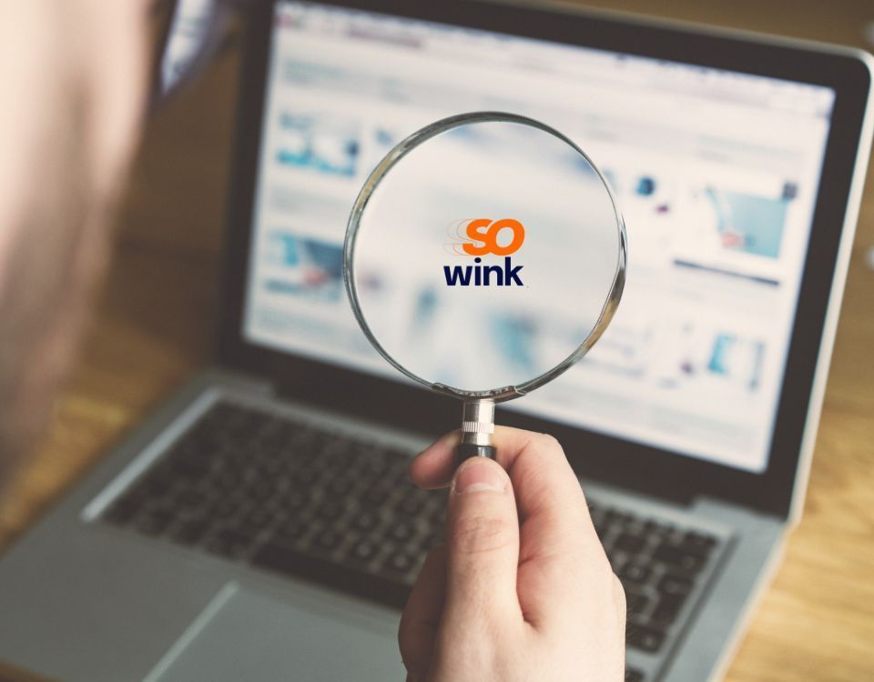 Une main tenant une loupe au-dessus de l'écran d'un ordinateur portable laisse apparaître le logo « so wink ».