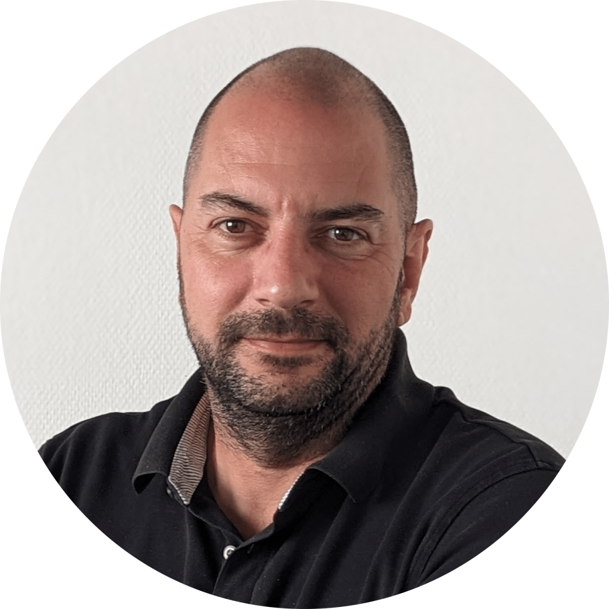 Une personne au crâne rasé et à la barbe fournie, portant un polo noir, se détache sur un fond blanc uni.