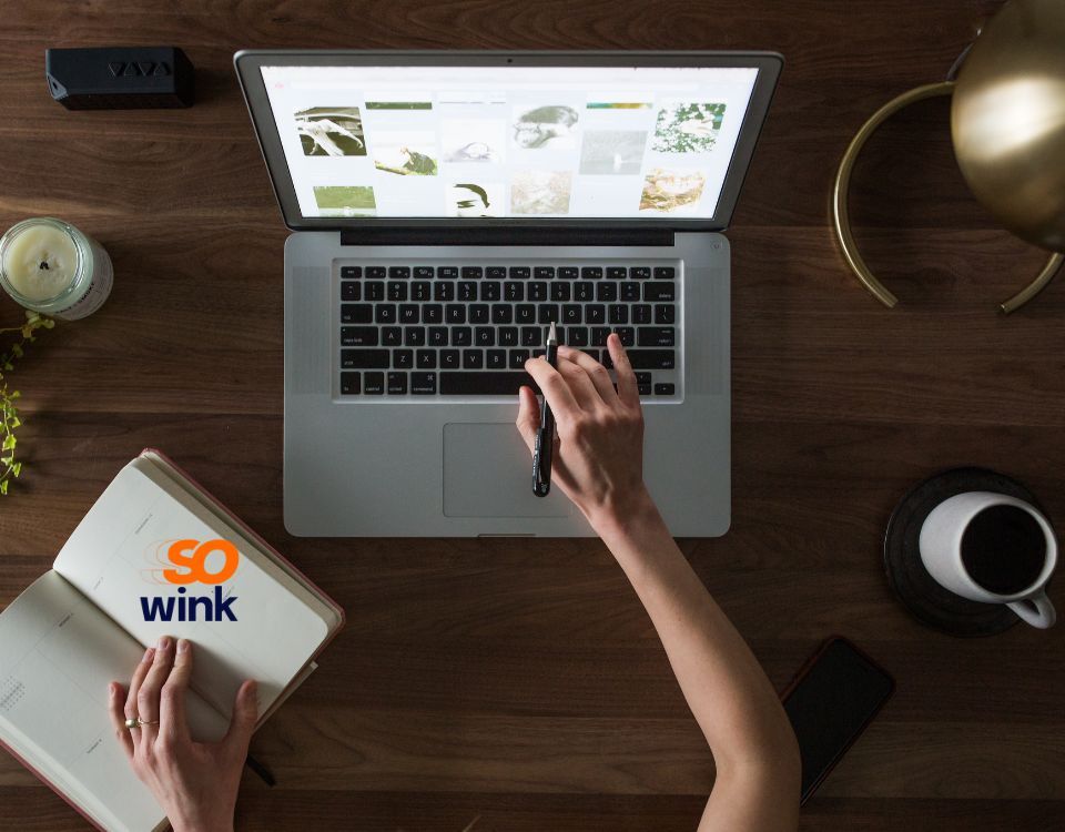 Vue de dessus d'une personne utilisant un ordinateur portable sur un bureau en bois avec une tasse de café et un carnet portant la marque « so wink ».