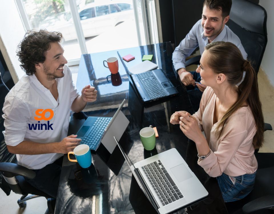 Trois personnes assises autour d'une table avec des ordinateurs portables, souriantes et discutant dans un bureau.