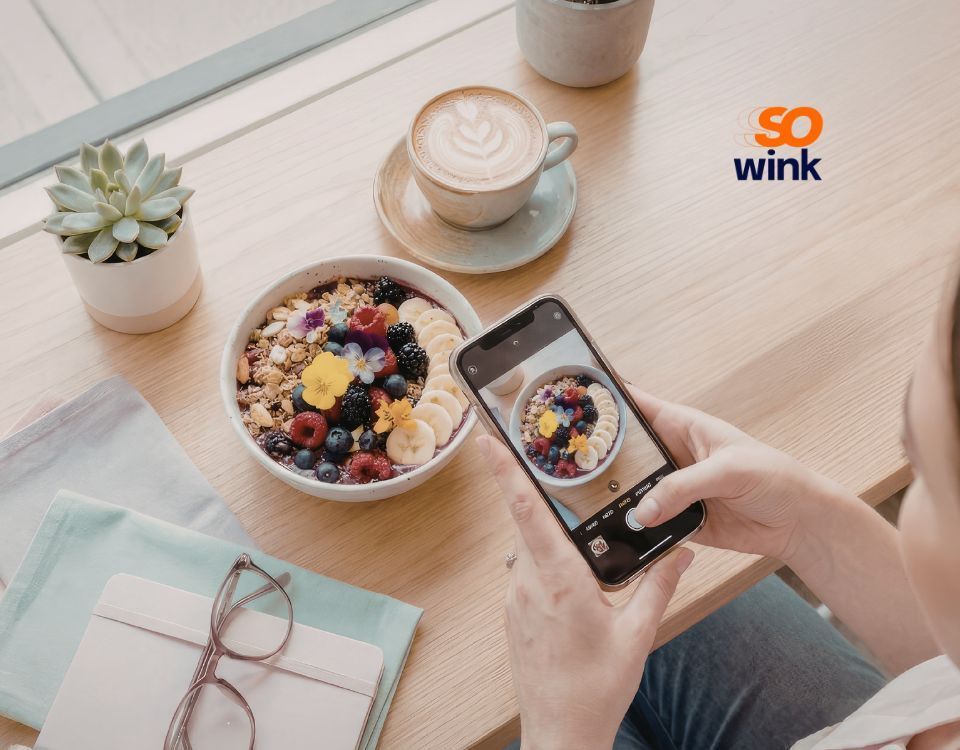 Une personne prend en photo un bol d'açaï avec son smartphone, assise à une table où se trouvent un café, une petite plante et un carnet.