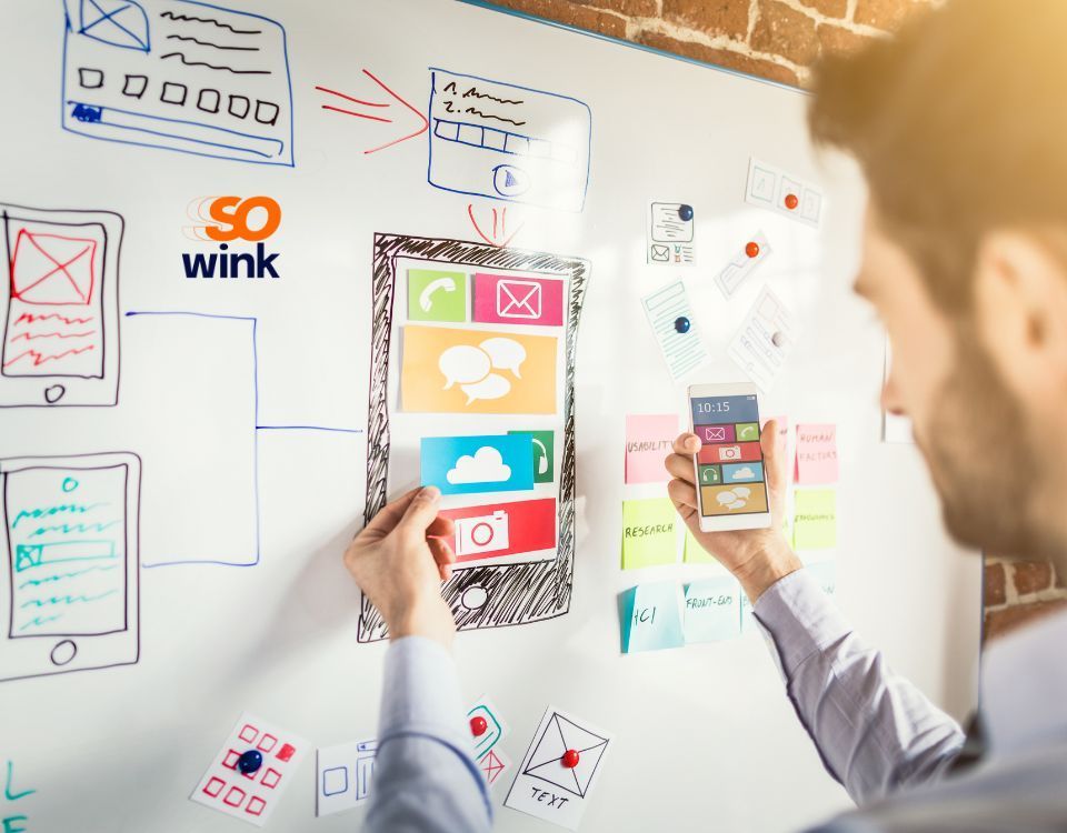 Une personne travaille sur un tableau blanc, y épinglant des croquis de conception d'interface utilisateur et utilisant un smartphone pour modèle.
