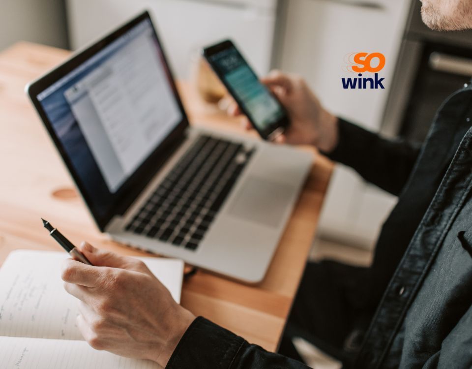 Une personne assise à un bureau, un ordinateur portable ouvert, tenant un stylo au-dessus d'un cahier tout en regardant un smartphone.