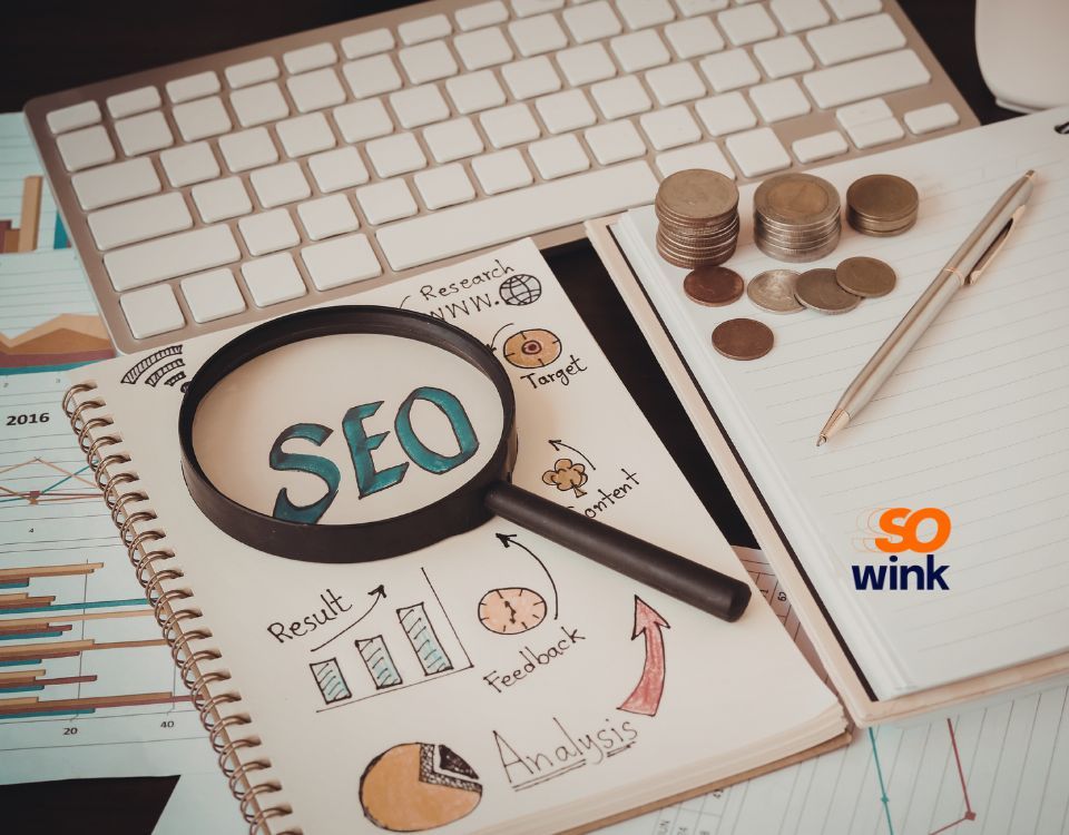 Une loupe au-dessus du mot « SEO » sur un bloc-notes avec des graphiques, des pièces de monnaie et un stylo posé sur un bureau.