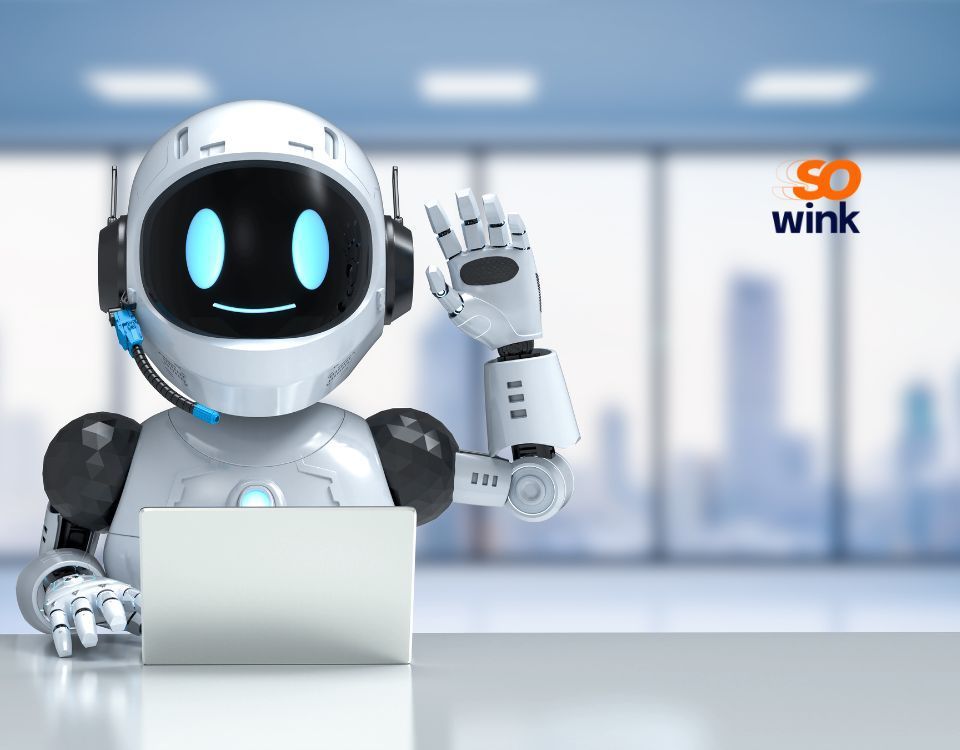 Robot blanc avec casque audio, faisant un signe de la main et souriant. Il travaille sur un ordinateur portable dans un bureau. Le logo « So Wink » est visible.