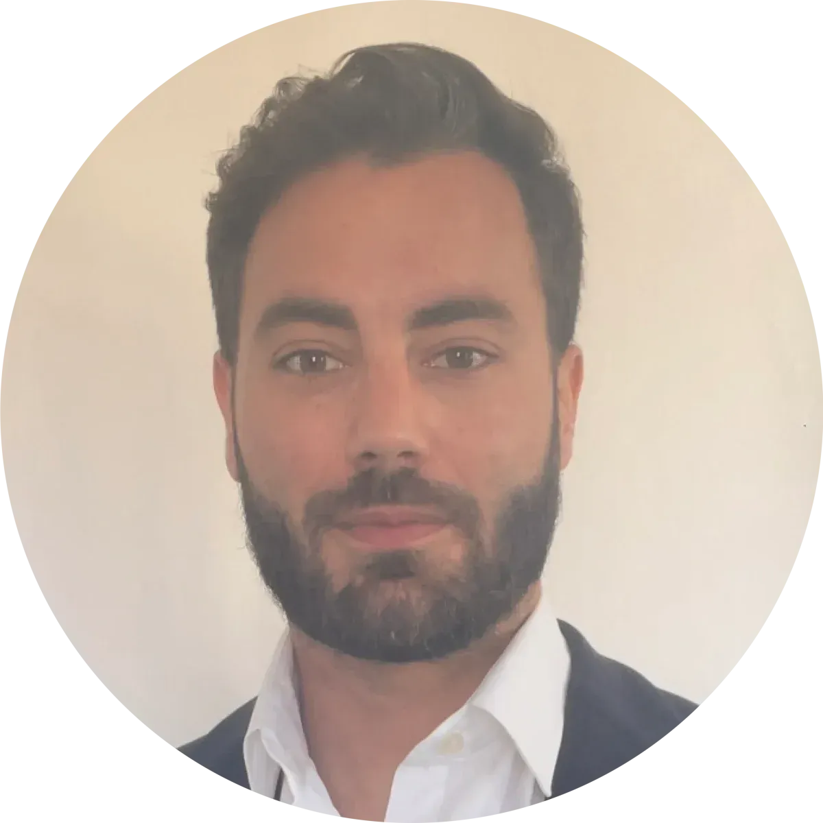 Un homme aux cheveux courts et foncés et à la barbe, portant une chemise blanche et une veste sombre, se détache sur un fond clair uni.