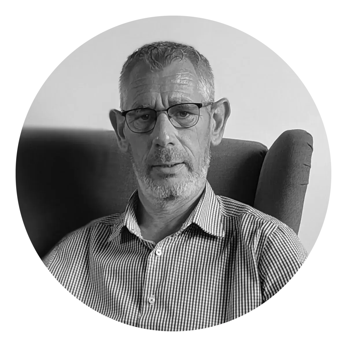 Portrait en noir et blanc d'une personne aux cheveux courts et portant des lunettes, vêtue d'une chemise à col et assise sur une chaise.