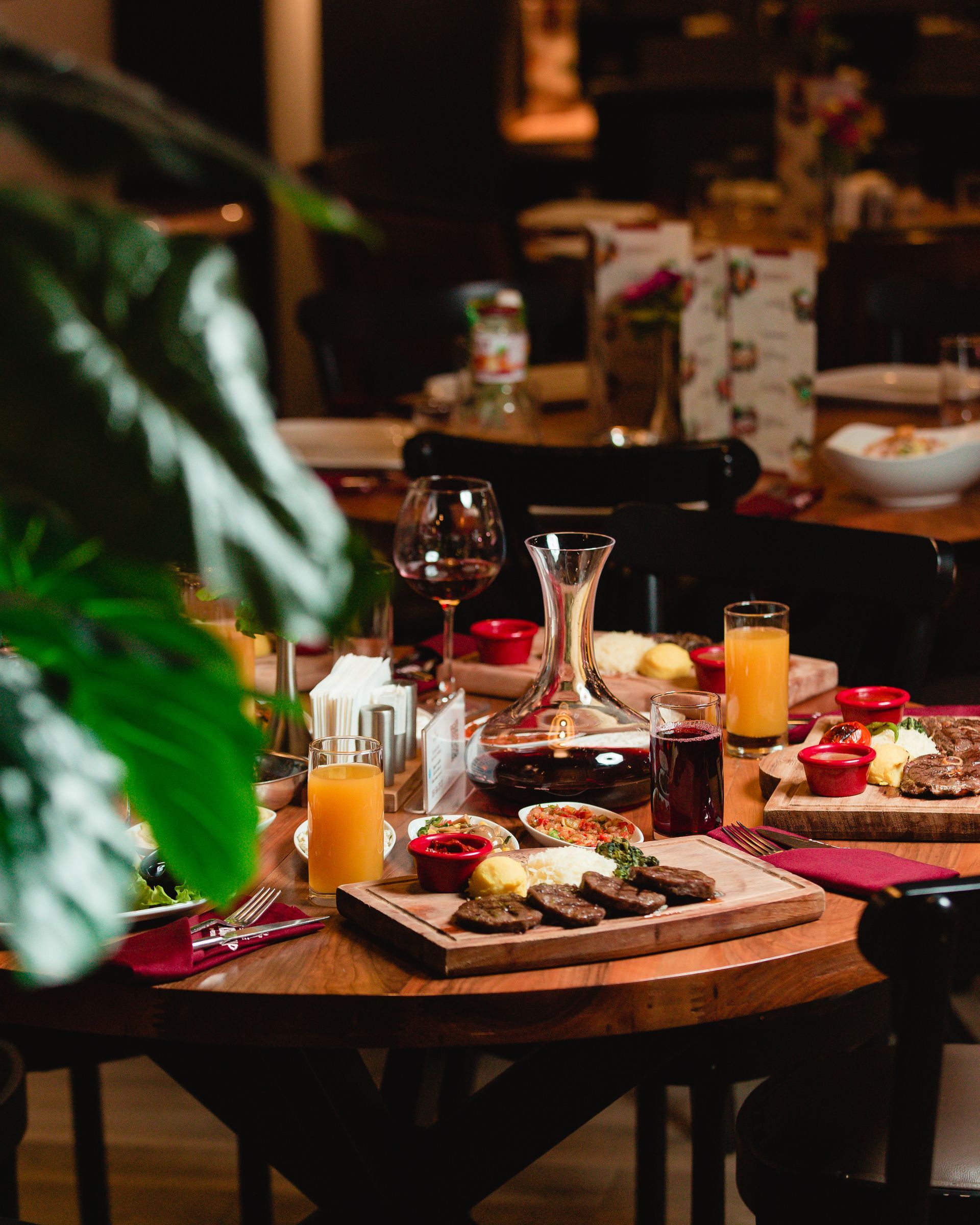 Une table en bois dressée pour un repas au restaurant, avec des plateaux en bois garnis de viandes grillées, de verres de jus de fruits et de vin.