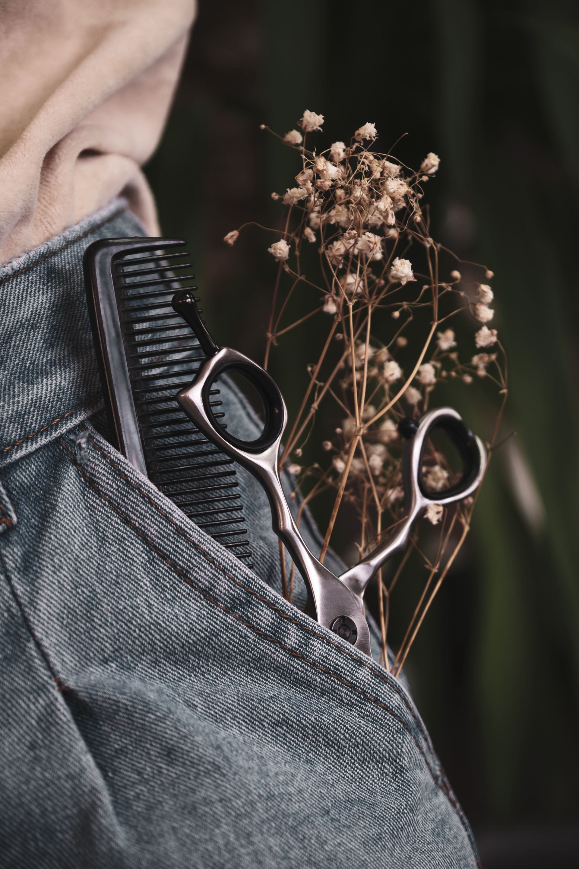 Des ciseaux de coiffure et un peigne noir glissés dans une poche en jean, à côté d'un brin de gypsophile séchée.