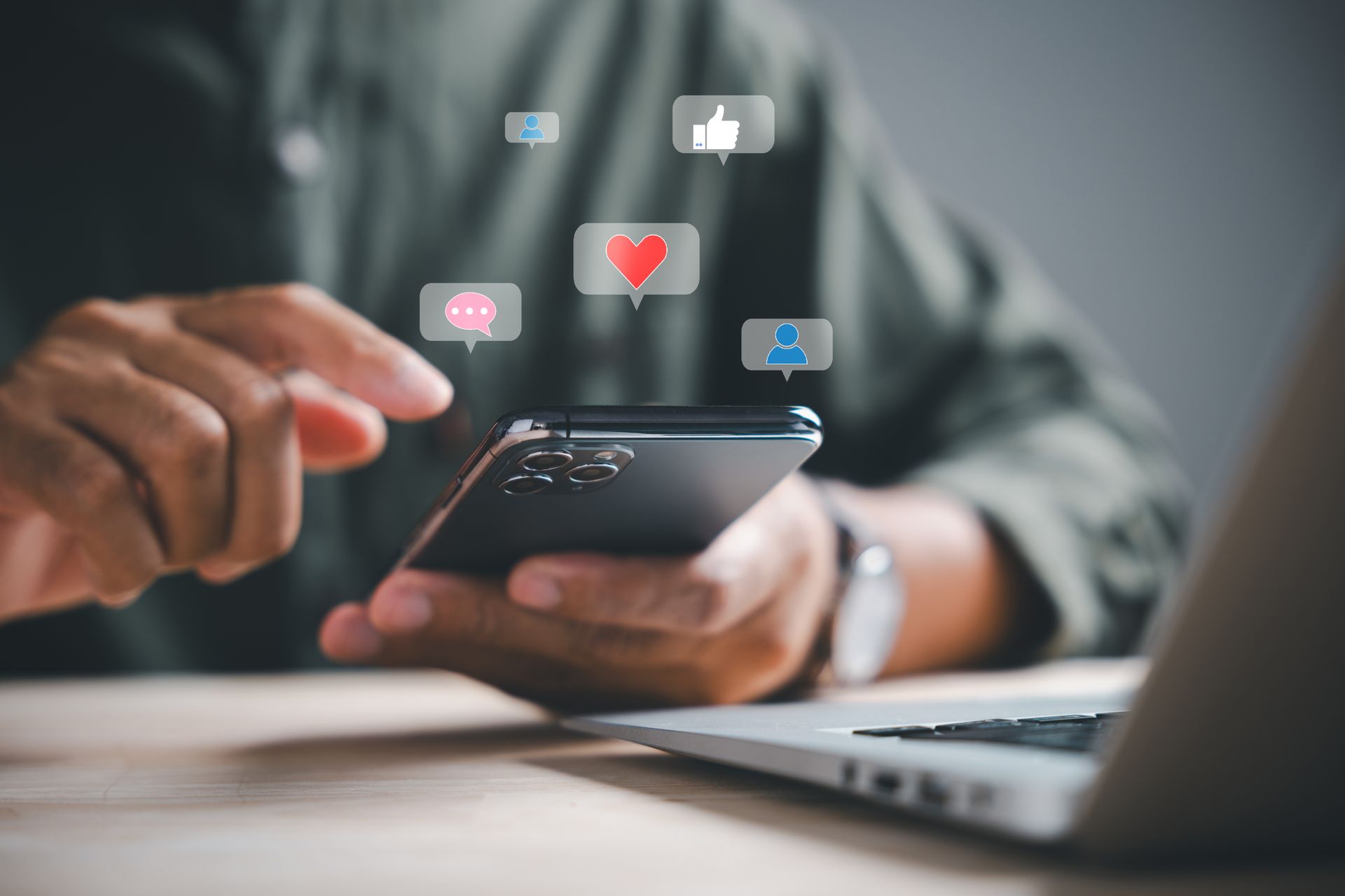 Une personne utilise un smartphone, entouré d'icônes de réactions de réseaux sociaux flottantes, avec un ordinateur portable visible à proximité.
