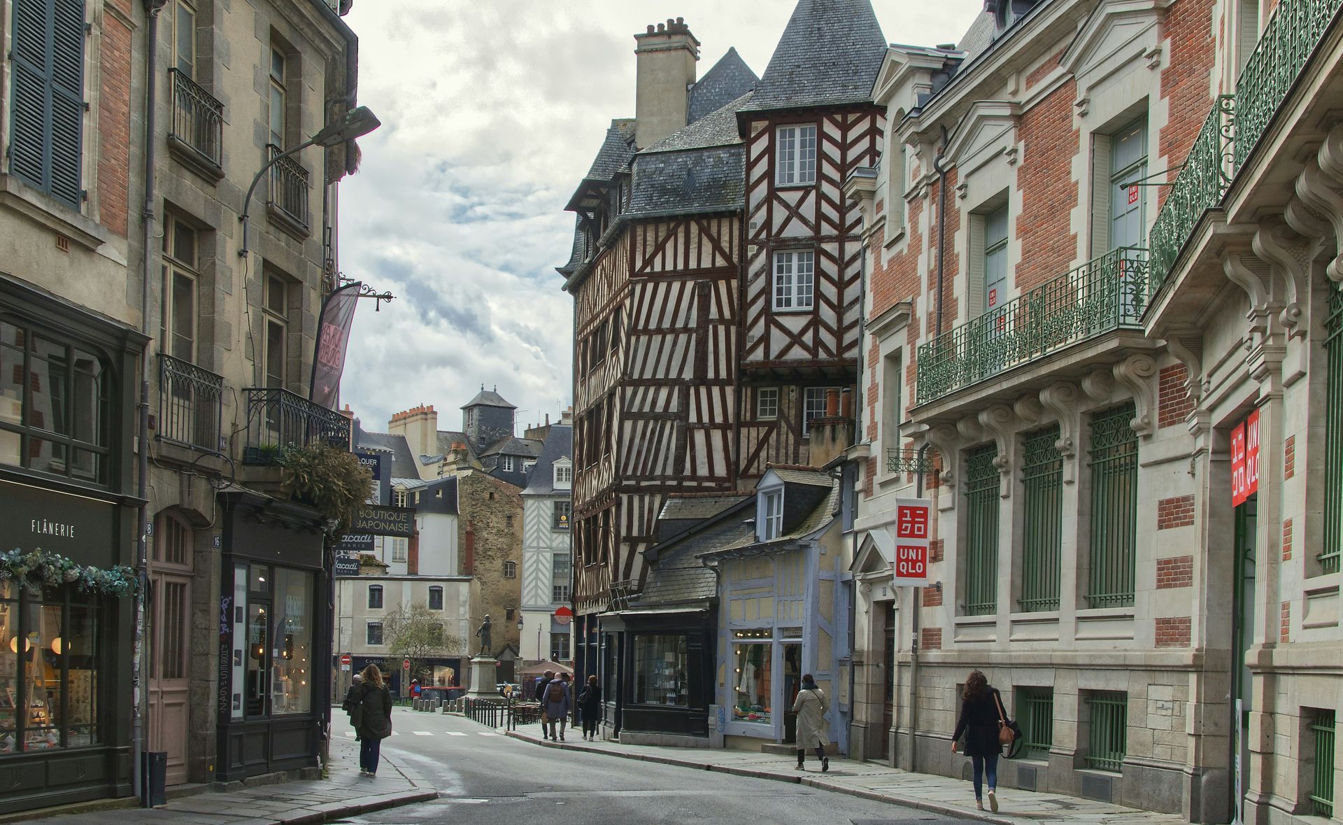 Une rue européenne étroite bordée de maisons traditionnelles à colombages et d'édifices en pierre, avec quelques piétons.