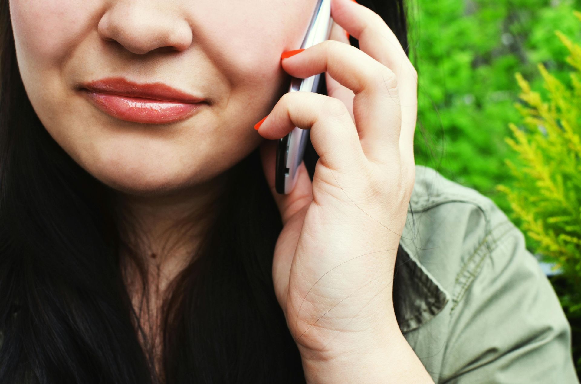 Une personne aux cheveux foncés parle au téléphone portable argenté devant un fond de verdure luxuriante.