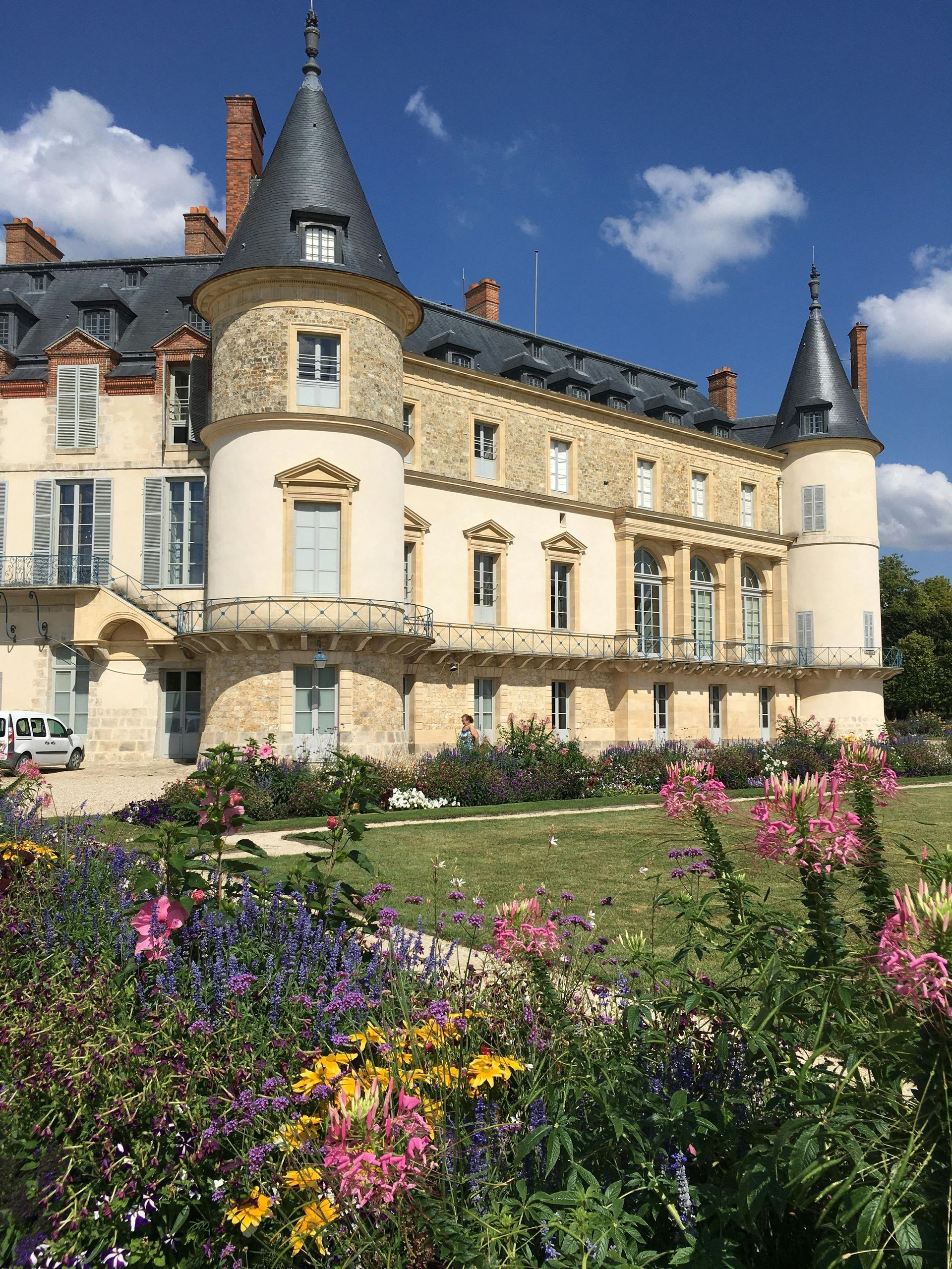 Un château de pierre, flanqué de deux tours d'angle proéminentes, se dresse derrière un jardin fleuri et coloré, sous un ciel d'un bleu éclatant.