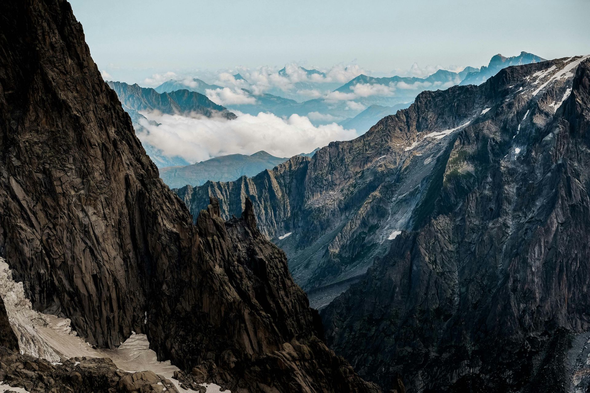 Des sommets montagneux escarpés et sombres encadrent une vallée profonde emplie de nuages ​​bas sous un ciel bleu pâle.