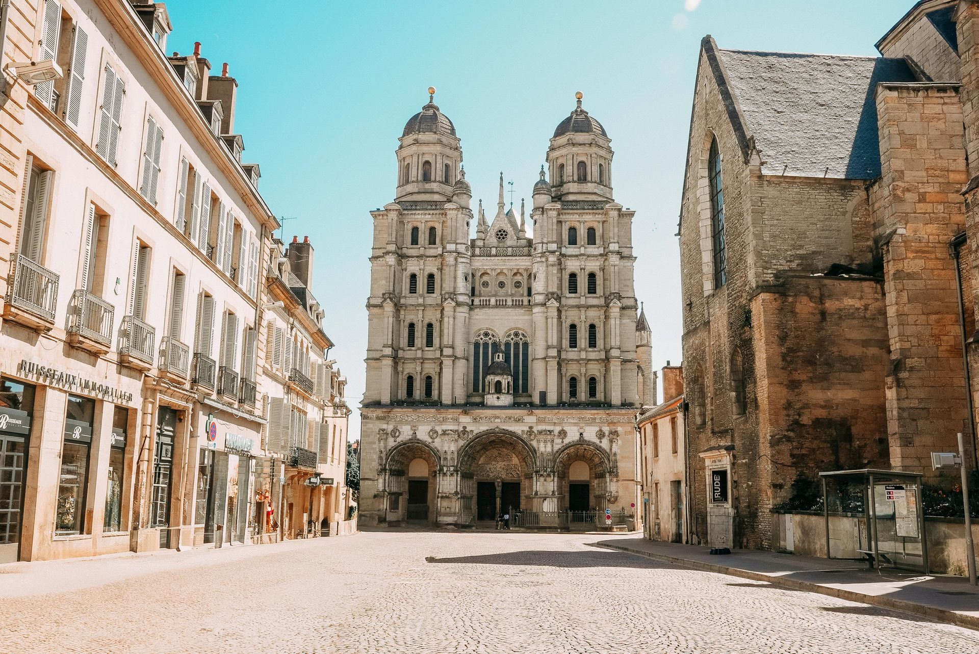 Sous un ciel bleu limpide, une rue mène à la façade ornée à deux tours de l'église Saint-Michel à Dijon, en France.