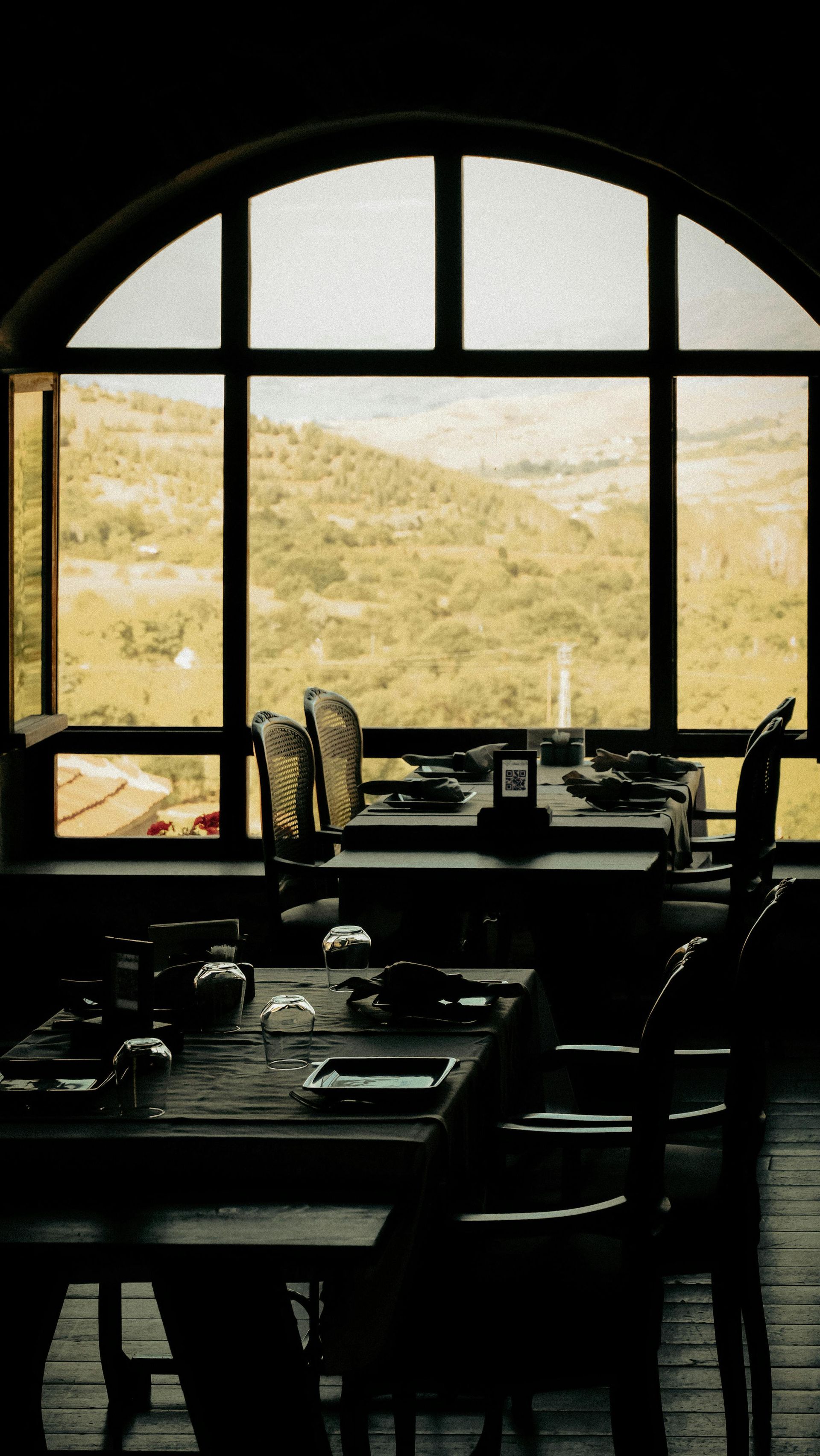Une salle à manger de restaurant avec des tables et des chaises en bois sombre disposées devant une grande fenêtre cintrée donnant sur une colline.