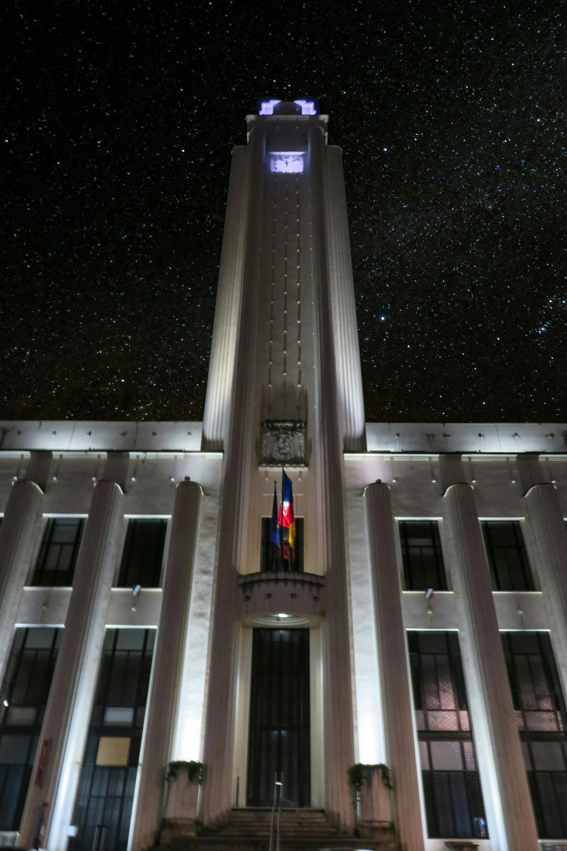 Un bâtiment en pierre illuminé, surmonté d'une tour d'horloge centrale, se détache sur un ciel nocturne étoilé.
