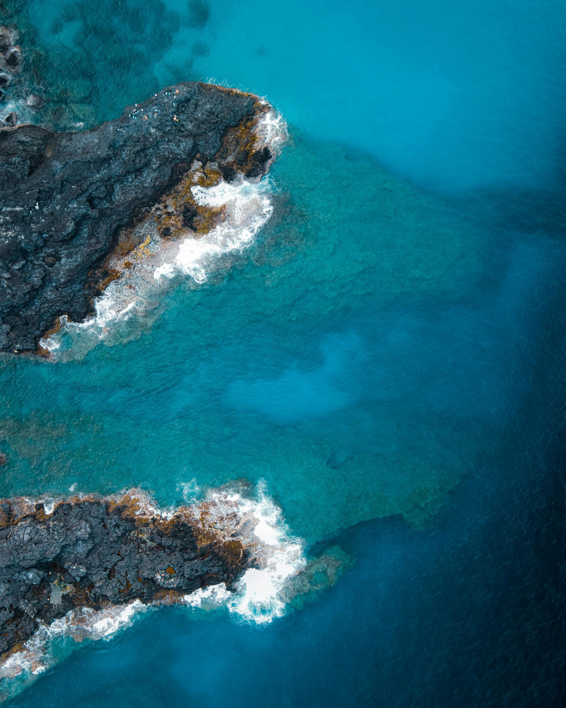 Vue aérienne de formations rocheuses volcaniques sombres émergeant des eaux turquoise de l'océan, contre lesquelles les vagues se brisent.