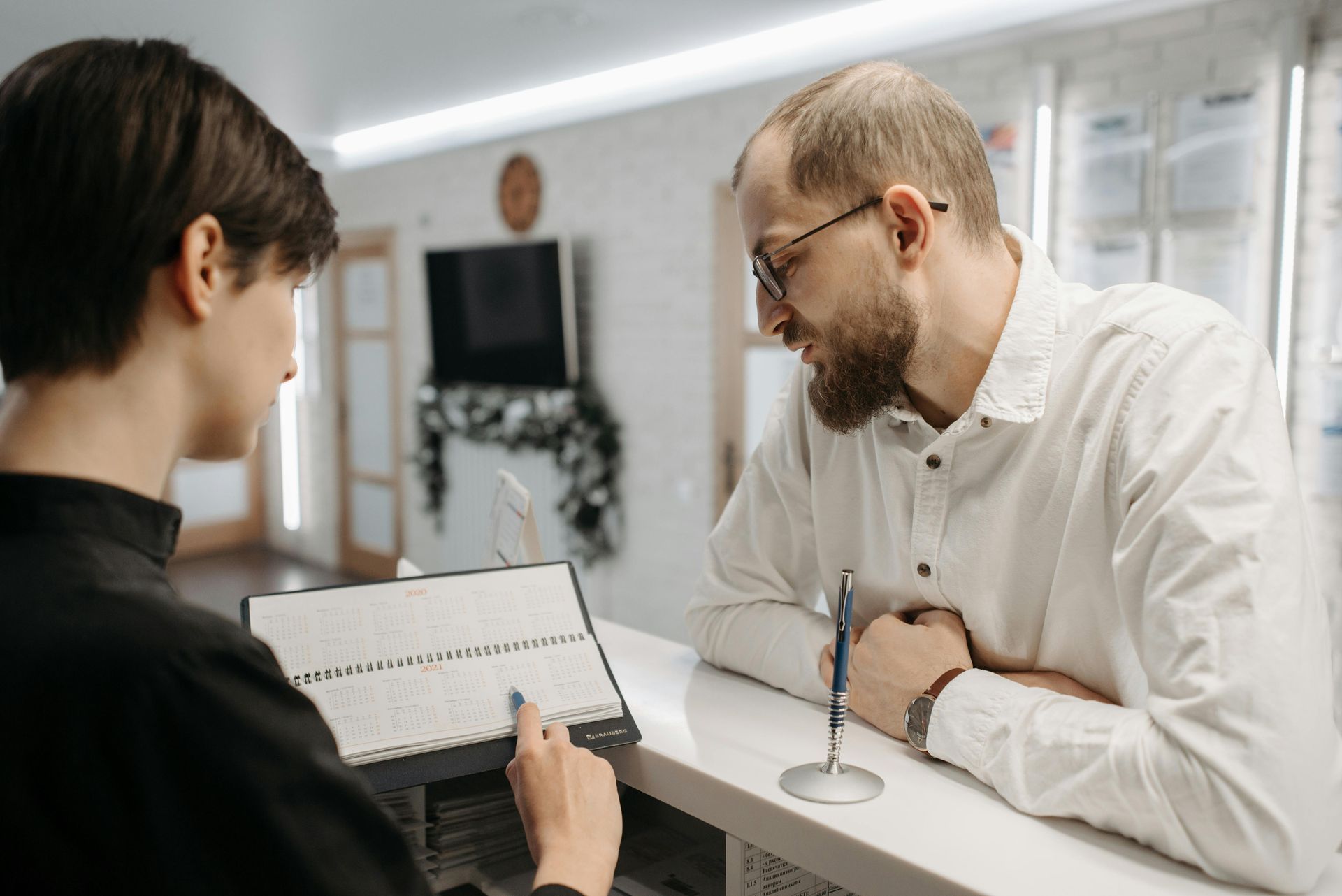 Une personne assise à un bureau montre un calendrier sur une tablette à un client qui se penche pour le regarder.