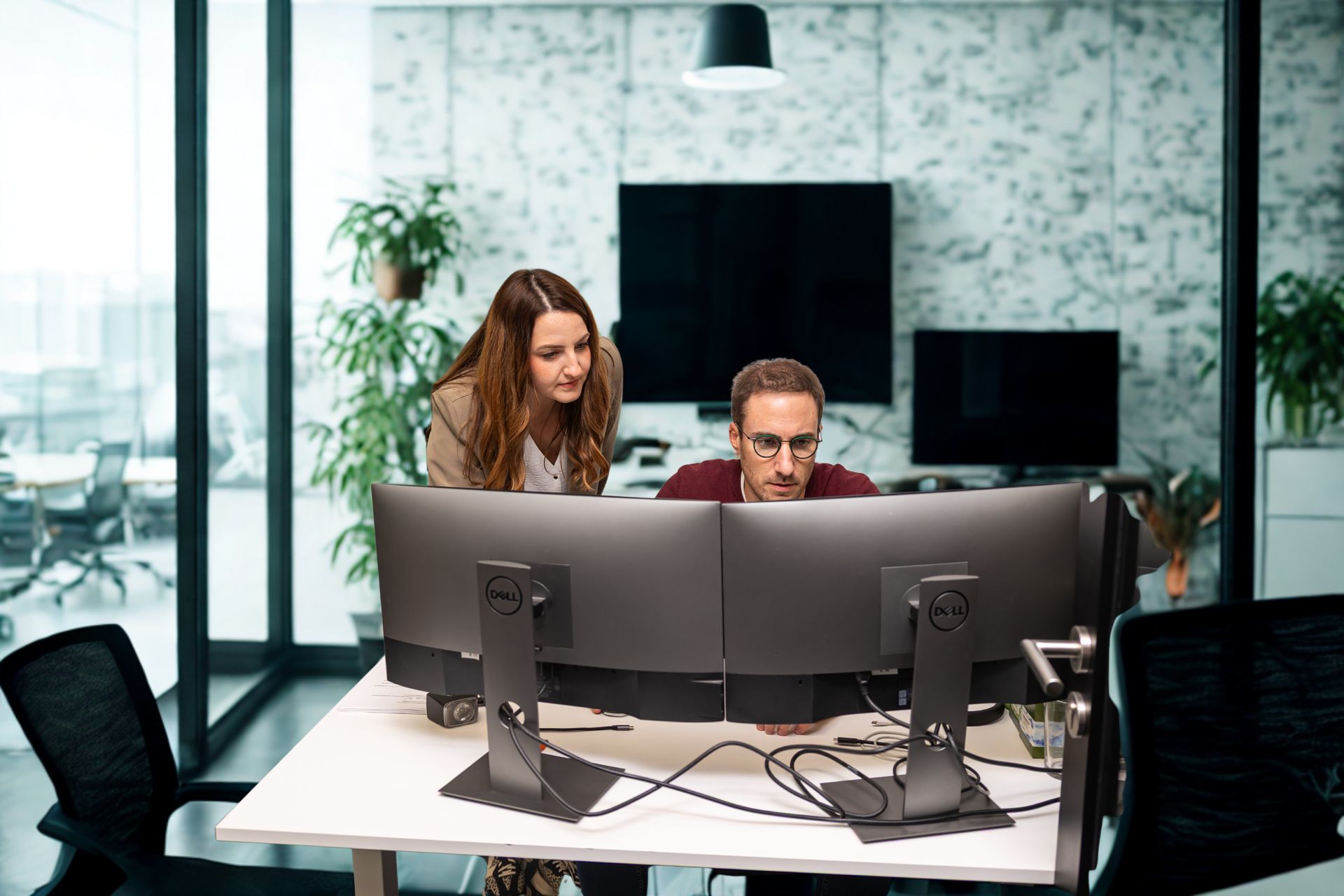 Deux collègues collaborent à un bureau équipé de deux écrans dans un bureau moderne aux parois de verre agrémenté de plantes.