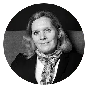 Portrait d'une personne aux cheveux mi-longs, portant un blazer foncé et une écharpe à motifs, sur fond sombre.