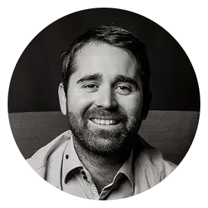 Portrait d'une personne souriante et barbue, portant une chemise boutonnée, sur fond sombre, dans un cadre circulaire.