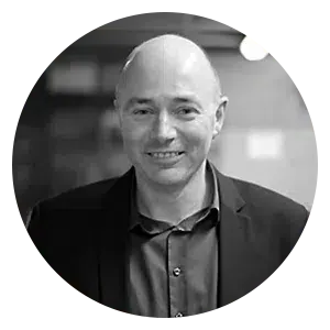 Portrait d'un homme souriant, vêtu d'un blazer sombre et d'une chemise boutonnée, encadré dans un cadre circulaire sur un fond flou.