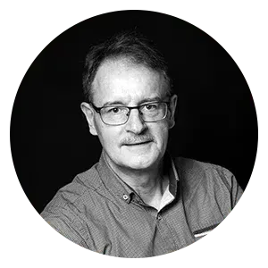 Portrait en noir et blanc d'une personne portant des lunettes et une chemise à col, centré sur un fond noir uni.