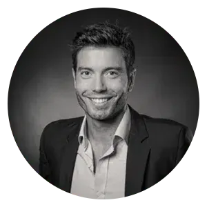 Portrait d'une personne souriante portant une chemise à col et un blazer sur un fond sombre et neutre.