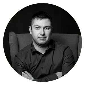 Portrait en noir et blanc d'une personne aux cheveux courts, portant une chemise boutonnée, assise dans un fauteuil, les bras croisés.