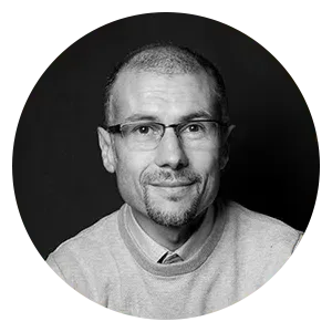 Portrait d'une personne aux cheveux courts, portant des lunettes et un bouc, vêtue d'un pull sur fond noir.