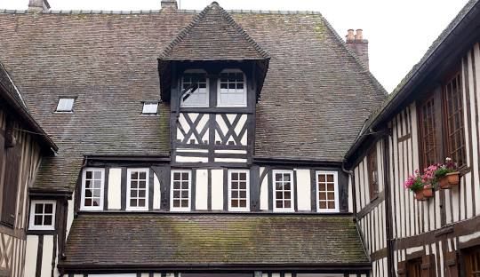 Un bâtiment à colombages avec des poutres en bois sombre, un remplissage blanc, un toit en bardeaux et une lucarne centrale à pignon.