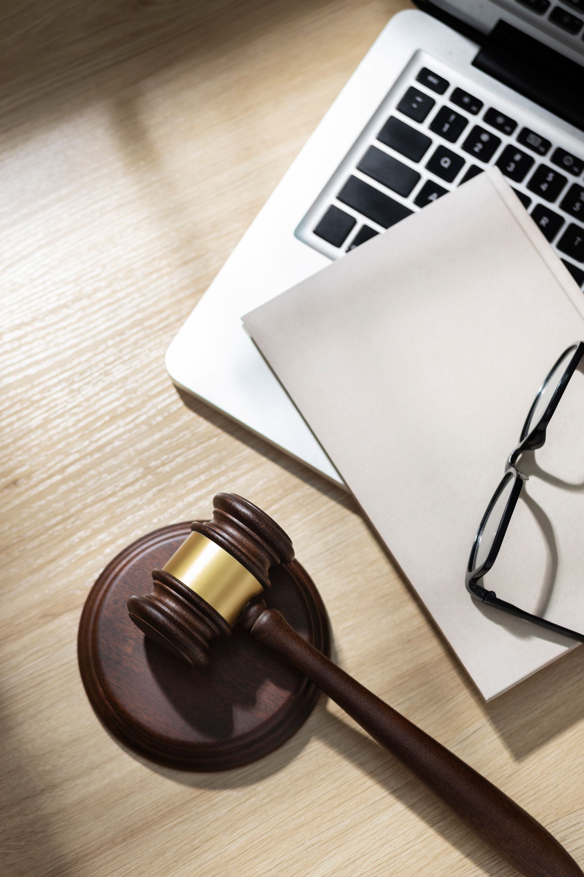 Un maillet en bois posé sur un bloc de frappe, à côté d'un cahier, d'une paire de lunettes et d'un ordinateur portable ouvert, sur un bureau en bois.
