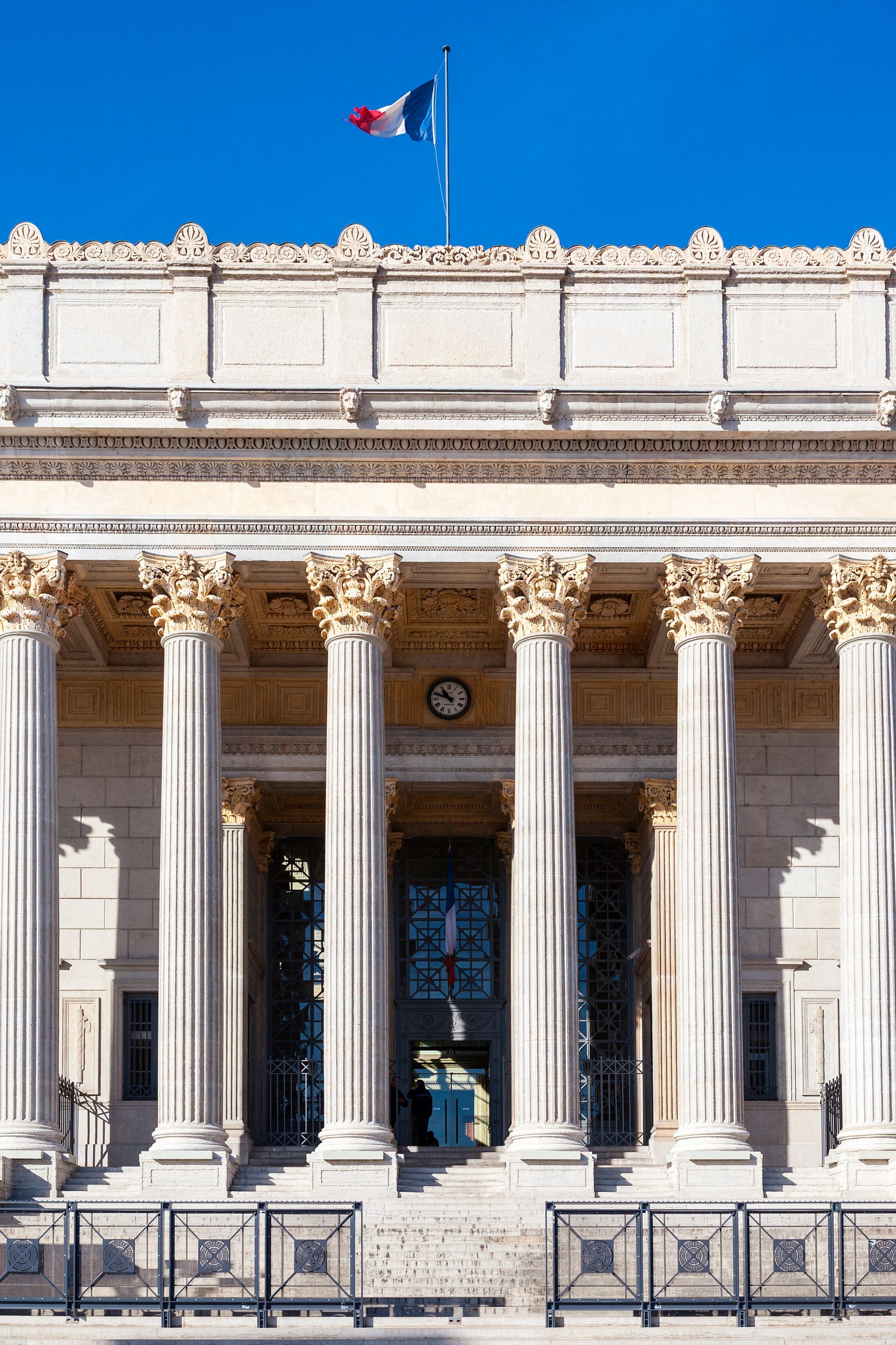 La façade d'un bâtiment néoclassique en pierre, ornée de hautes colonnes et surmontée d'un drapeau français flottant sur un ciel bleu.