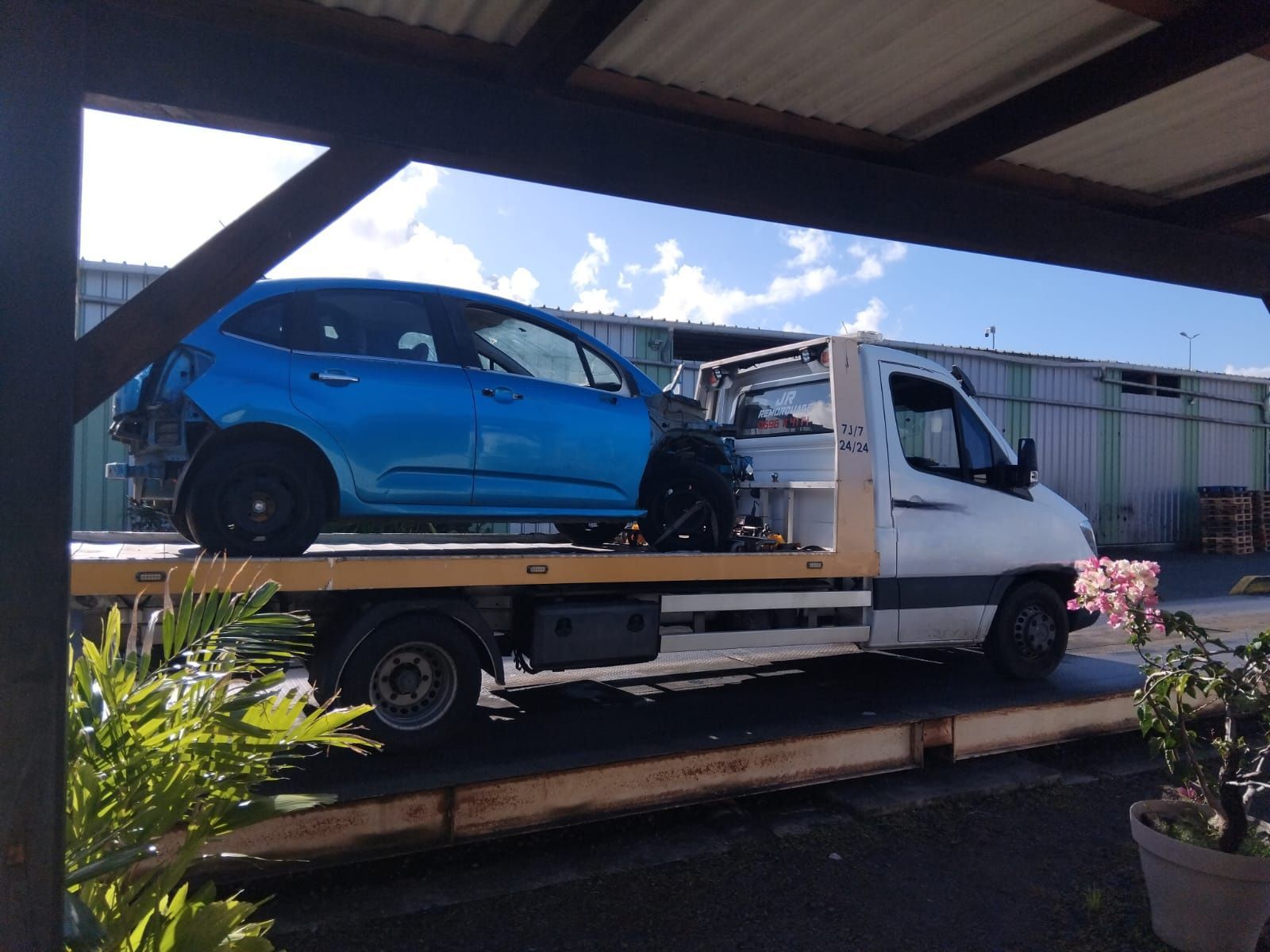 Une voiture à hayon chargée sur une dépanneuse à plateau sous une allée couverte.