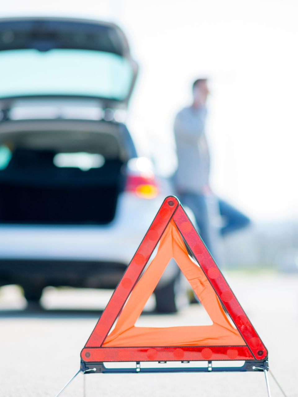 Triangle de signalisation rouge devant une voiture en panne. Une personne parle au téléphone.
