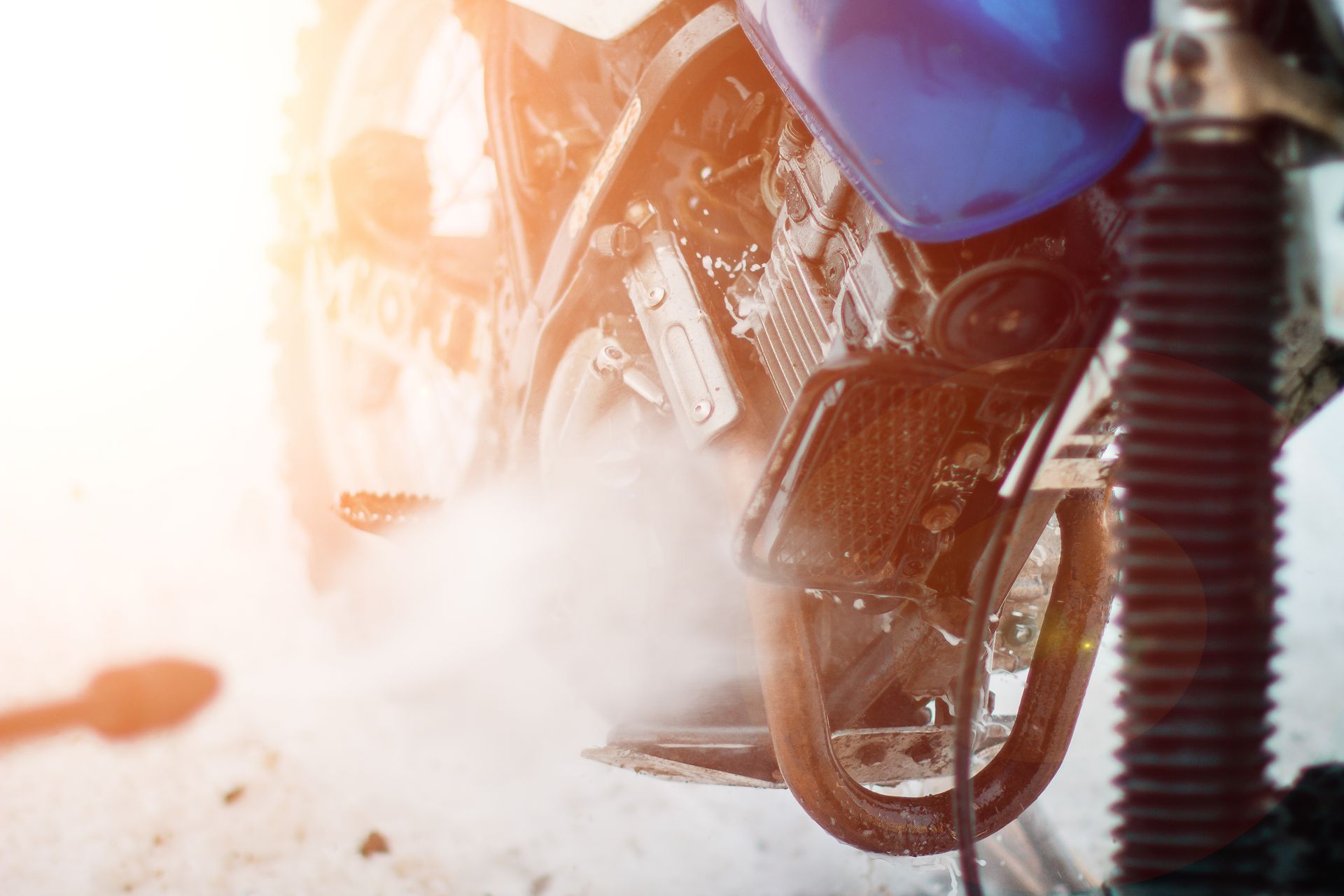 La roue d'une moto soulève de la poussière sur une route, un motard en tenue est visible.