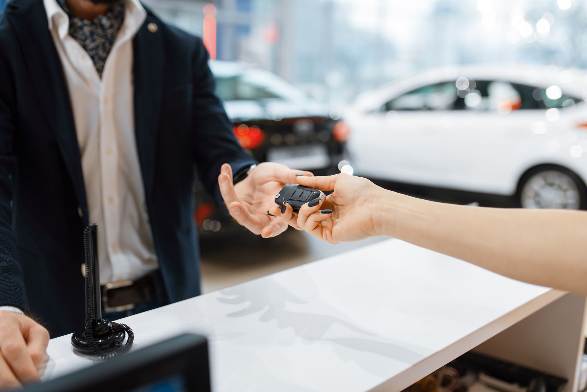 Des mains échangent des clés de voiture au comptoir d'une concession automobile, une voiture étant visible en arrière-plan.