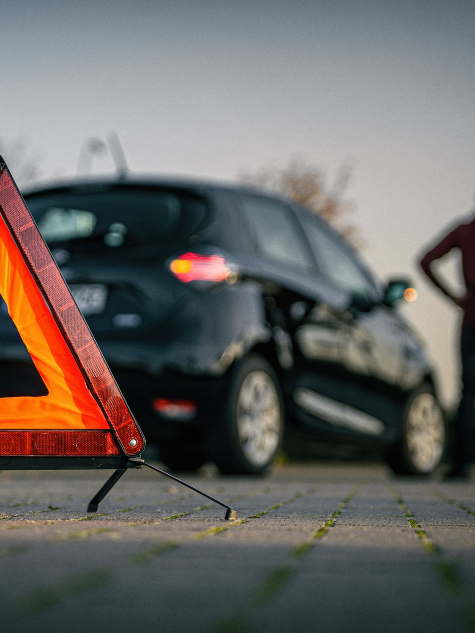 Triangle de signalisation devant une voiture noire en panne, une personne se tient à proximité.