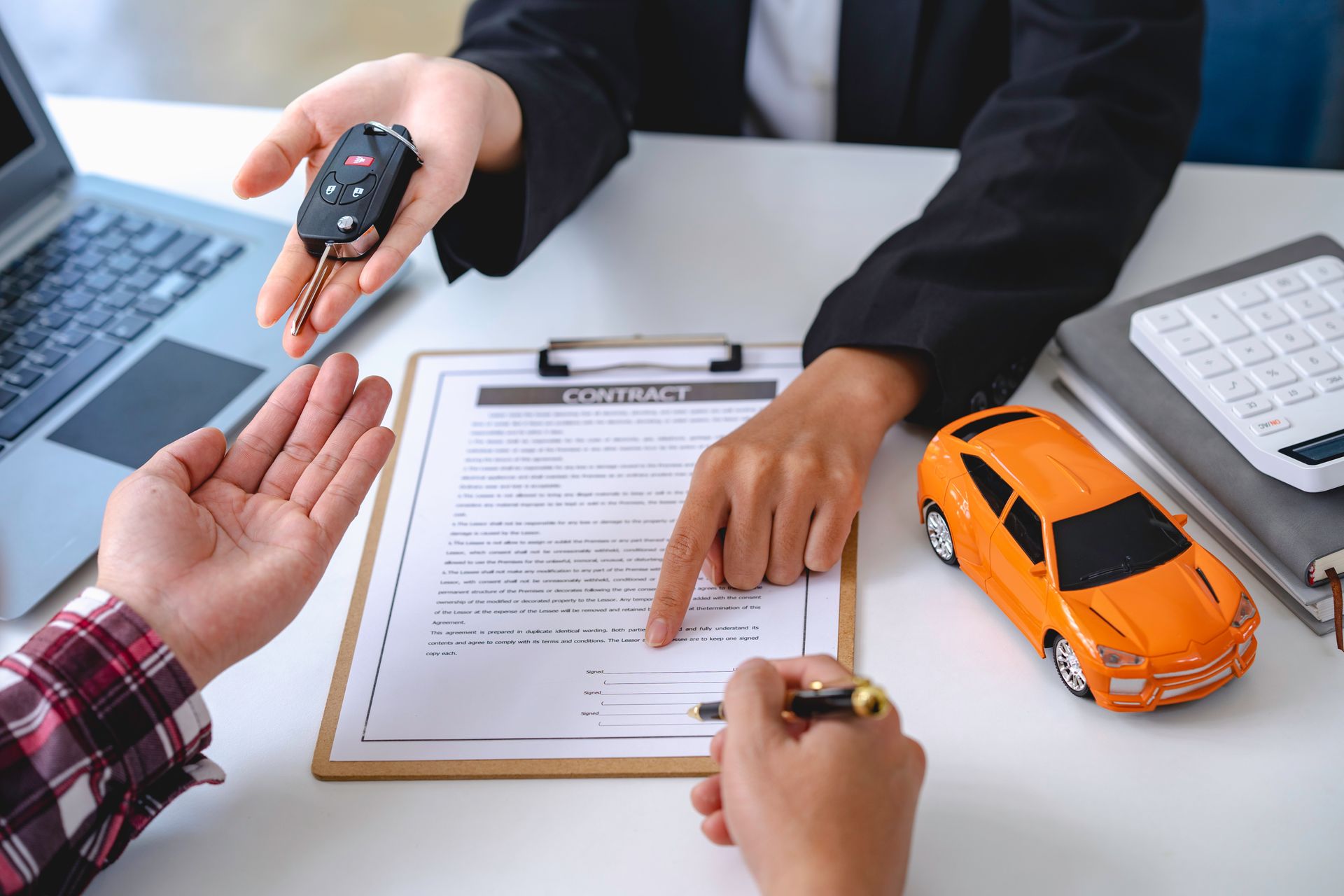 Des mains s'échangent des clés de voiture au-dessus de papiers, à côté d'une petite voiture et d'un ordinateur portable posés sur un bureau.