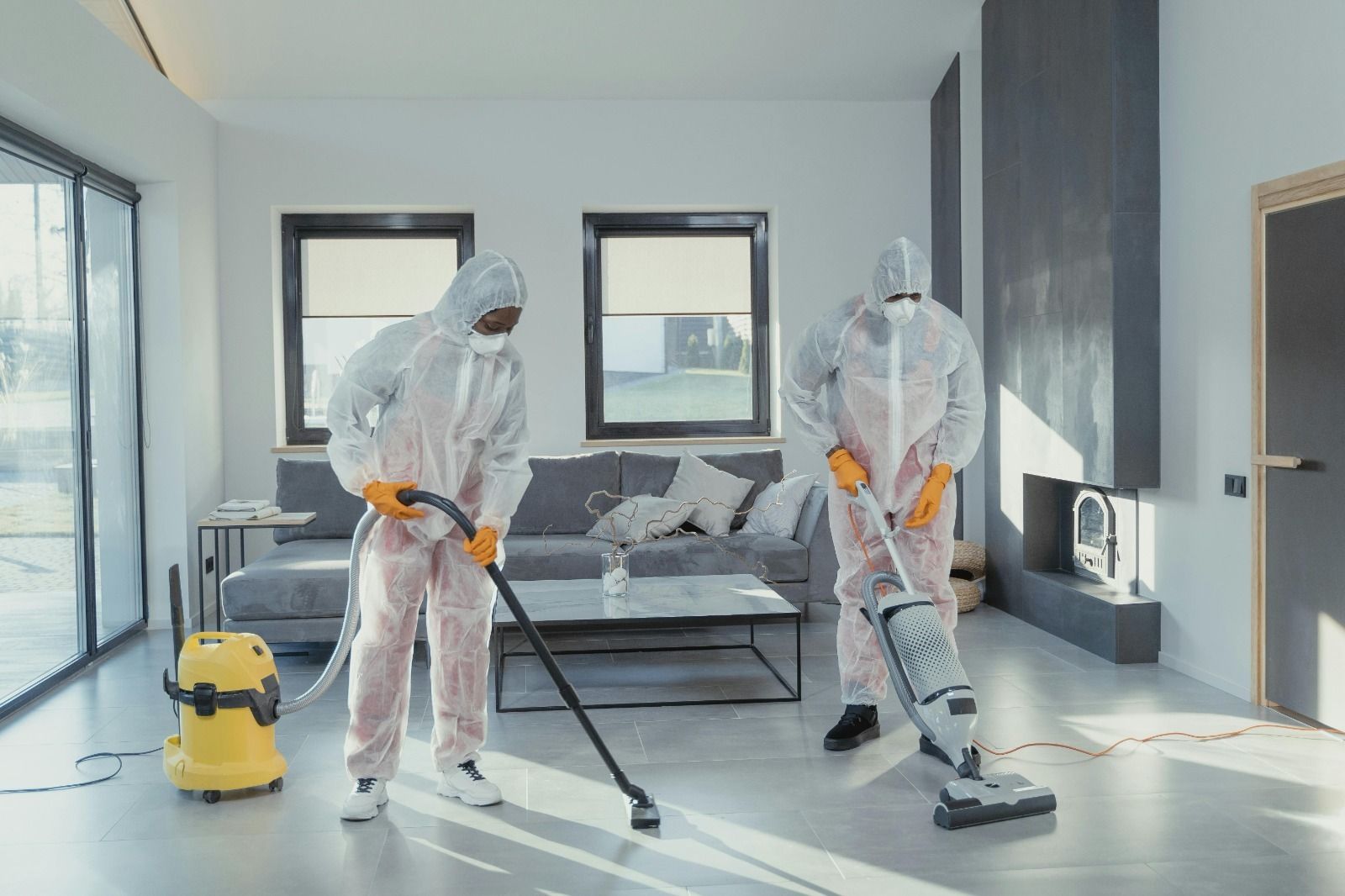 Dos hombres con trajes de protección están limpiando una sala de estar con aspiradoras.