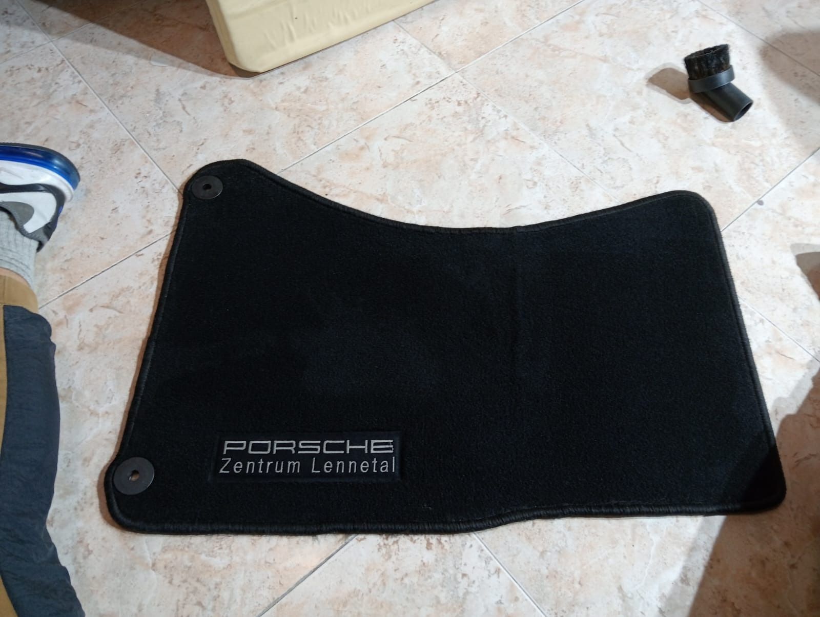 Una alfombrilla Porsche negra reposa sobre un suelo de baldosas al lado de un par de zapatos.