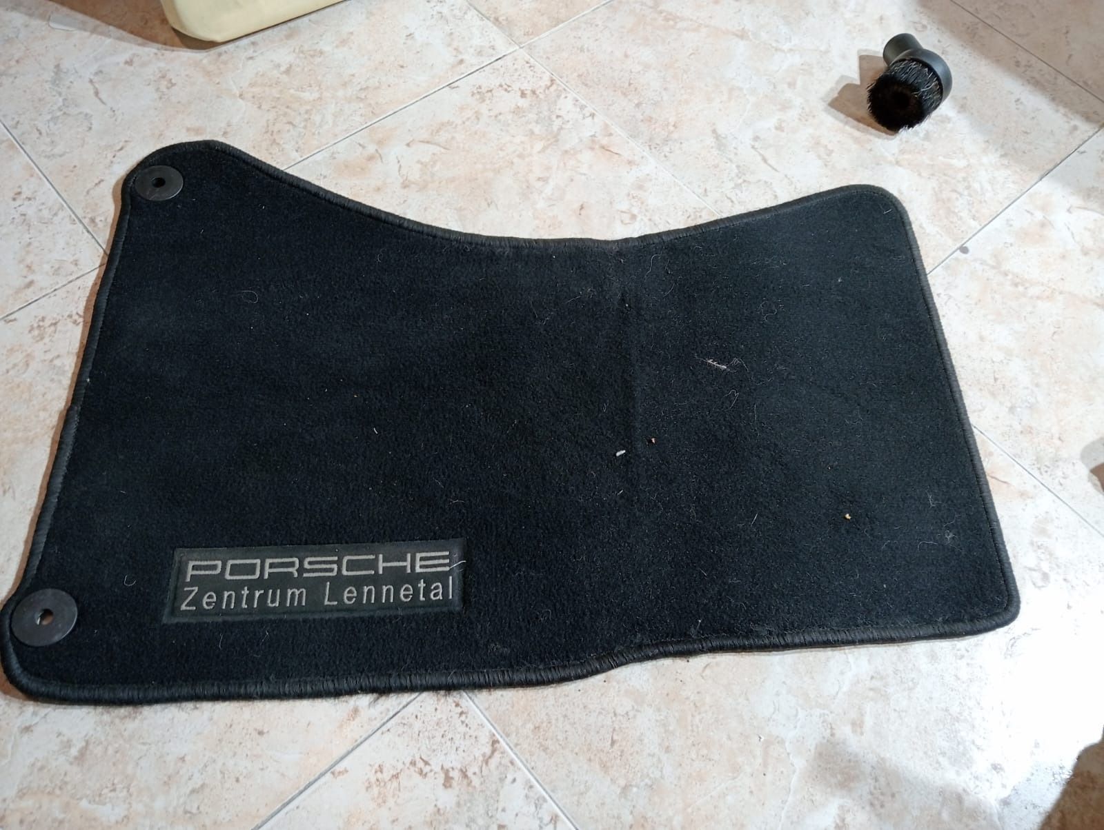 Una alfombrilla de asiento de Porsche de color negro se encuentra sobre un suelo de baldosas.